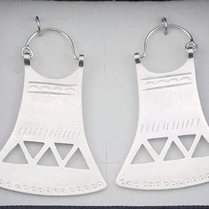 May include: A pair of silver hoop earrings with a geometric design. The earrings are made of metal and have a triangular shape with cutouts. The earrings are approximately 5 cm in diameter.