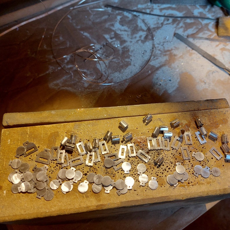 May include: A close-up of a wooden surface with a variety of small metal pieces. The pieces are in different shapes and sizes, including circles, squares, and rectangles. Some of the pieces have a shiny finish, while others have a matte finish.