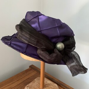 May include: A purple satin hat with a black ribbon and a silver bead accent. The hat is on a wooden hat stand.
