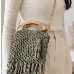 May include: A sage green macrame shoulder bag with a wooden handle and gold chain strap. The bag features a tassel and fringe detail. The bag is approximately 10 inches wide and 12 inches tall.