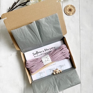 May include: A kit for making a macrame sunflower wall hanging. The kit includes pink macrame cord, wooden beads, and instructions. The kit is packaged in a brown cardboard box with tissue paper.
