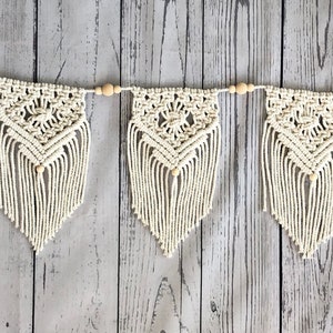 May include: A white macrame banner with wooden beads strung between each triangle-shaped piece. The banner has a fringe detail on the bottom of each piece.