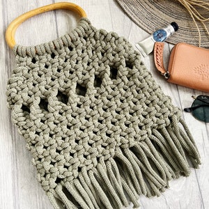 K&ouml;nnte beinhalten: Eine gr&uuml;ne geh&auml;kelte Tasche mit einem Holzgriff und Fransen. Die Tasche ist mit einem Makramee-Stich gearbeitet und hat ein einzigartiges durchbrochenes Design.