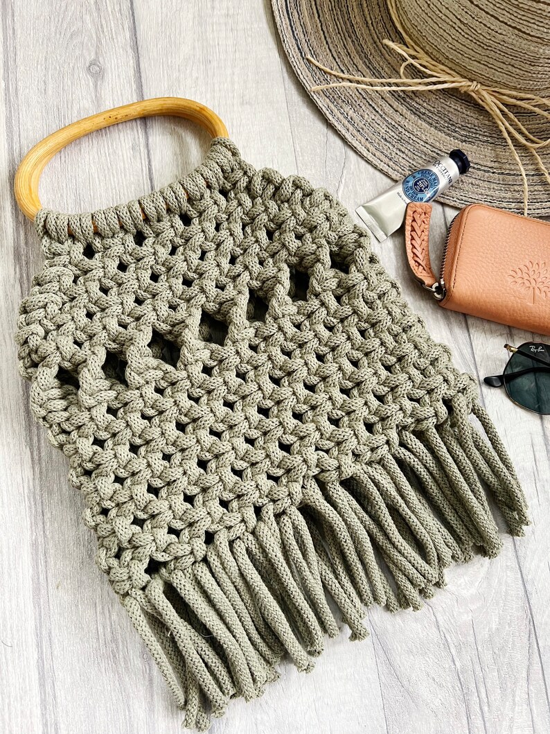 K&ouml;nnte beinhalten: Eine gr&uuml;ne geh&auml;kelte Tasche mit einem Holzgriff und Fransen. Die Tasche ist mit einem Makramee-Stich gearbeitet und hat ein einzigartiges durchbrochenes Design.