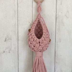 May include: A pink macrame plant hanger with a wooden ring at the top and a long tassel. The hanger is hanging on a white wooden wall.