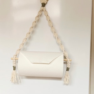 May include: A white paper towel holder made of macrame rope with wooden dowels and tassels. The holder is hanging from a hook on a white wall.