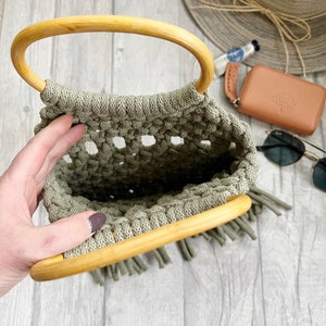 K&ouml;nnte beinhalten: Eine hellgr&uuml;ne geh&auml;kelte Tasche mit einem Holzgriff und Fransen. Die Tasche ist offen und leer.