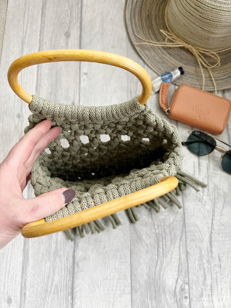 K&ouml;nnte beinhalten: Eine hellgr&uuml;ne geh&auml;kelte Tasche mit einem Holzgriff und Fransen. Die Tasche ist offen und leer.