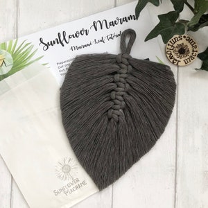 May include: A grey macrame leaf hanging decoration with a loop at the top. The leaf is made with a thick cord and has a fringed edge.