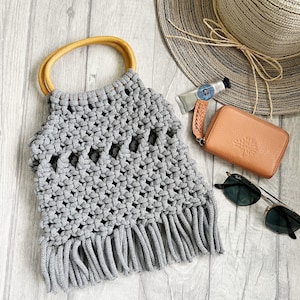 May include: A grey crocheted tote bag with a wooden handle and fringe detail. The bag is on a white wood background with a straw hat, sunglasses, and a small brown wallet.