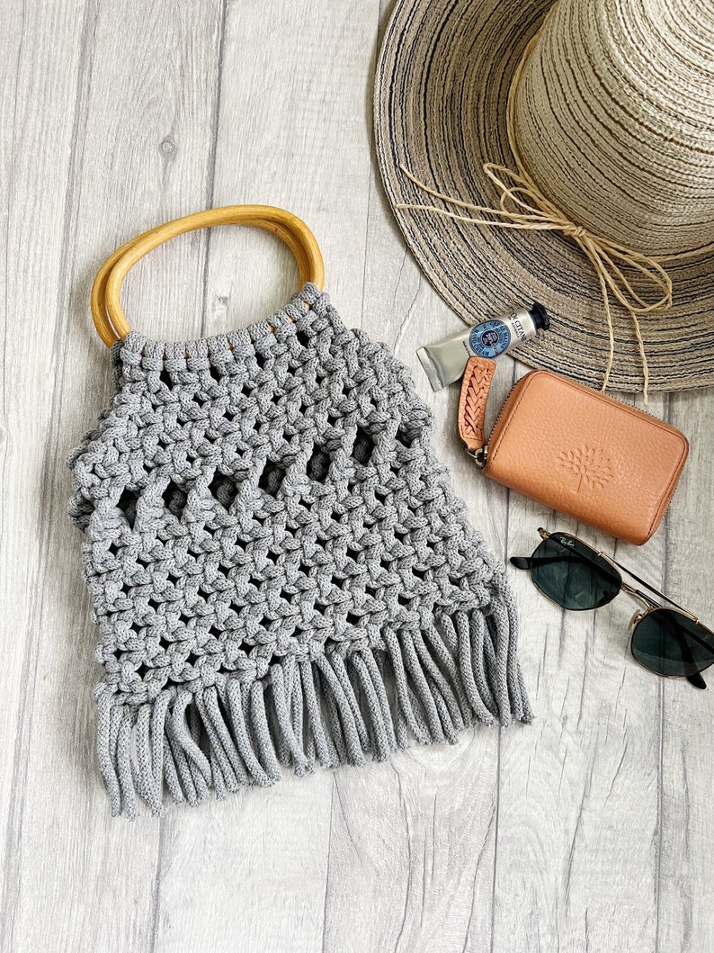 May include: A grey crocheted tote bag with a wooden handle and fringe detail. The bag is on a white wood background with a straw hat, sunglasses, and a small brown wallet.