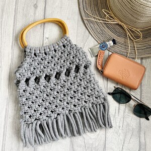 May include: A grey crocheted tote bag with a wooden handle and fringe detail. The bag is on a white wood background with a straw hat, sunglasses, and a small brown wallet.