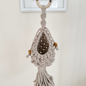 May include: A macrame plant hanger with a wooden ring at the top and a tassel at the bottom. The hanger is made of natural beige cotton cord and has small wooden beads.