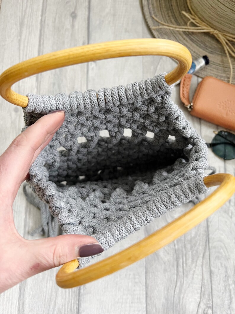 May include: A grey macrame bag with wooden handles. The bag is made using a knotting technique and has a fringed edge.