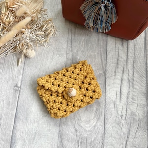 May include: A small mustard yellow crocheted pouch with a wooden button closure. The pouch is sitting on a light wood surface.