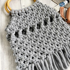 May include: Grey crocheted tote bag with wooden handles and fringe. The bag has a geometric pattern and is made with thick yarn.