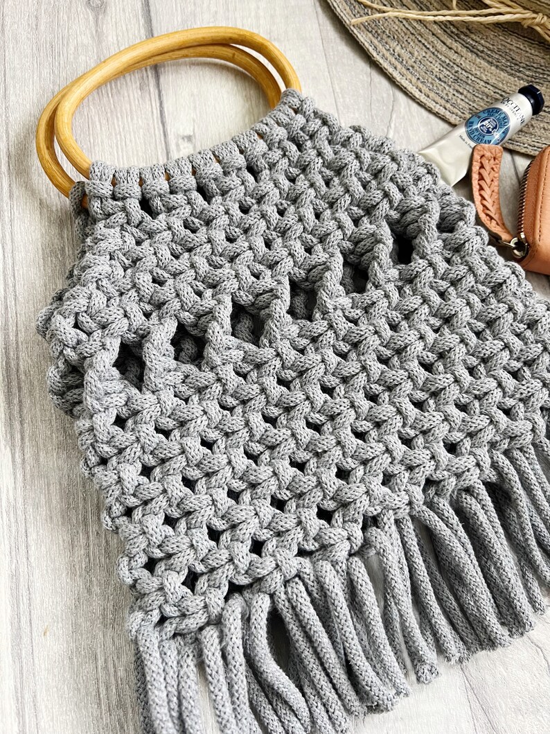 May include: Grey crocheted tote bag with wooden handles and fringe. The bag has a geometric pattern and is made with thick yarn.