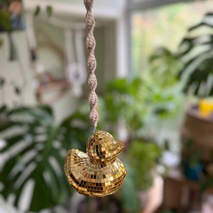 May include: A gold disco ball duck hanging from a macrame cord. The duck is made of small mirrored tiles.