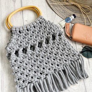 May include: Grey crocheted tote bag with a wooden handle and fringe detail. The bag is made with a thick, textured yarn and has a woven pattern.