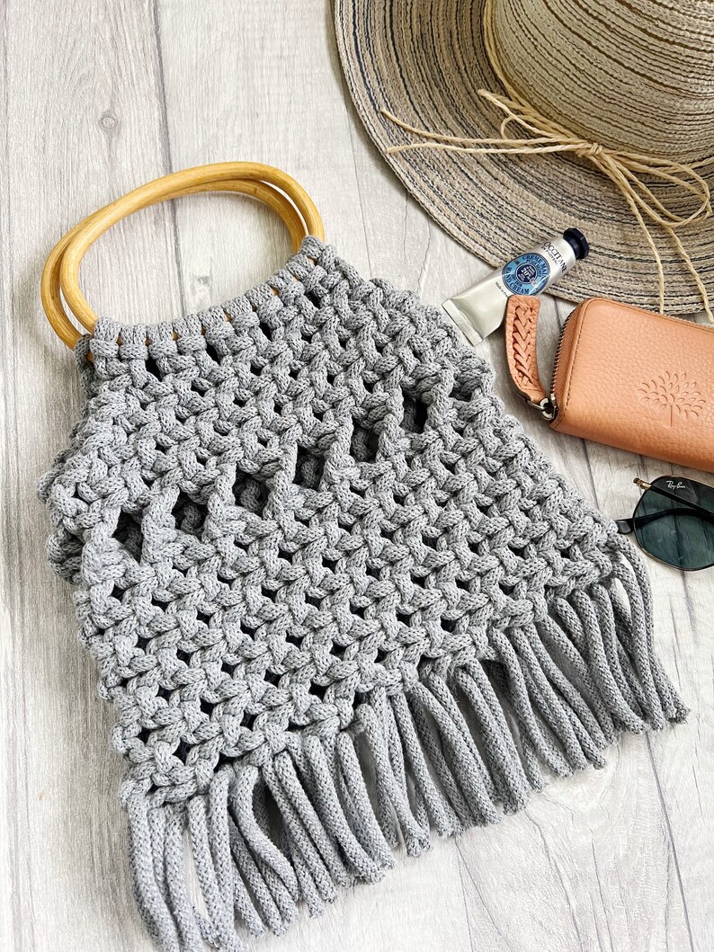May include: Grey crocheted tote bag with a wooden handle and fringe detail. The bag is made with a thick, textured yarn and has a woven pattern.