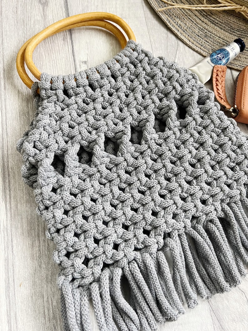 May include: Grey crocheted tote bag with wooden handles and fringe. The bag has a geometric pattern and is made with thick yarn.