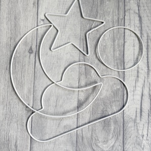 May include: White metal wireframe shapes of a star, moon, circle, and cloud. The celestial shapes are arranged on a gray wood-grain background. These shapes could be used for a nursery mobile or other craft projects.
