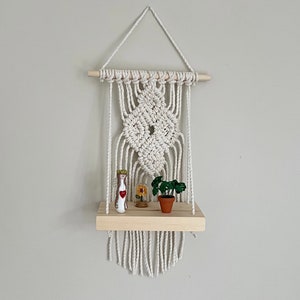 May include: A white macrame wall shelf with a wooden shelf and a small potted plant, a glass bottle with a figurine inside, and a small glass dome with a flower inside.
