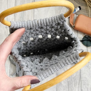May include: A grey macrame bag with wooden handles. The bag is made using a knotting technique and has a fringed bottom.