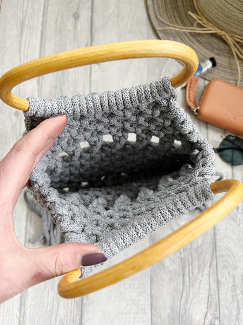 May include: A grey macrame bag with wooden handles. The bag is made using a knotting technique and has a fringed bottom.