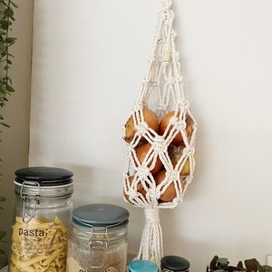 May include: A white macrame hanging onion holder with a wooden ring at the top. The holder is filled with six brown onions. There are several glass jars with labels on the counter below the holder, including one labelled "pasta" and another labelled "sugar".