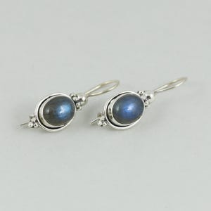 May include: A pair of silver drop earrings featuring oval-shaped, iridescent blue gemstones. The earrings have a simple, elegant design with a small decorative element near the ear wire. The gemstones have a subtle, shimmering effect.