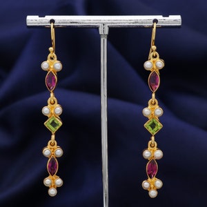 May include: Gold dangle earrings with green and pink gemstones and white pearls. The earrings are hanging from a silver stand.