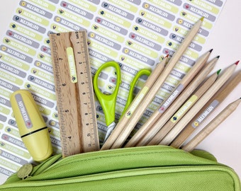 May include: A green pencil case containing wooden pencils, a wooden ruler, and green scissors. A sheet of rectangular adhesive labels with the name MAXIMILIAN and small monster illustrations is visible. Each item features a matching name label.