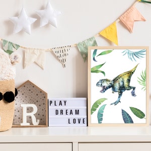 May include: A framed watercolor print of a green and yellow dinosaur with green leaves on a white background. The print is hanging on a white wall with a light up sign that says "PLAY DREAM LOVE" and a white stuffed animal in a basket.