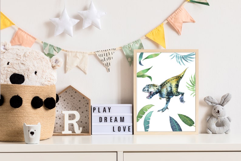 May include: A framed watercolor print of a green and yellow dinosaur with green leaves on a white background. The print is hanging on a white wall with a light up sign that says "PLAY DREAM LOVE" and a white stuffed animal in a basket.