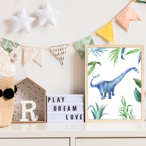 May include: A framed watercolor print of a blue dinosaur in a jungle setting. The print is surrounded by a light wood frame and is hanging on a white wall. The wall is decorated with a string of colorful flags.