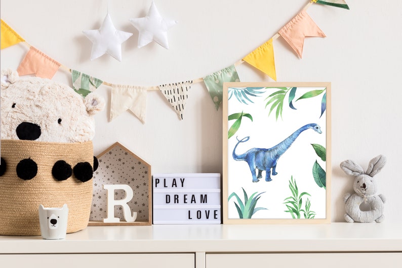 May include: A framed watercolor print of a blue dinosaur in a jungle setting. The print is surrounded by a light wood frame and is hanging on a white wall. The wall is decorated with a string of colorful flags.