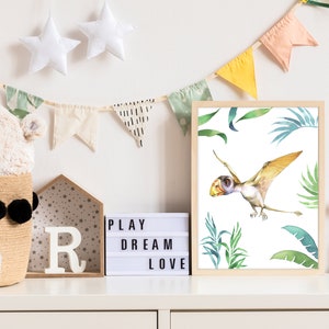 May include: A watercolor illustration of a pterodactyl flying in a jungle setting. The illustration is framed and hanging on a white wall with a light box that reads "PLAY DREAM LOVE" and a stuffed animal rabbit.