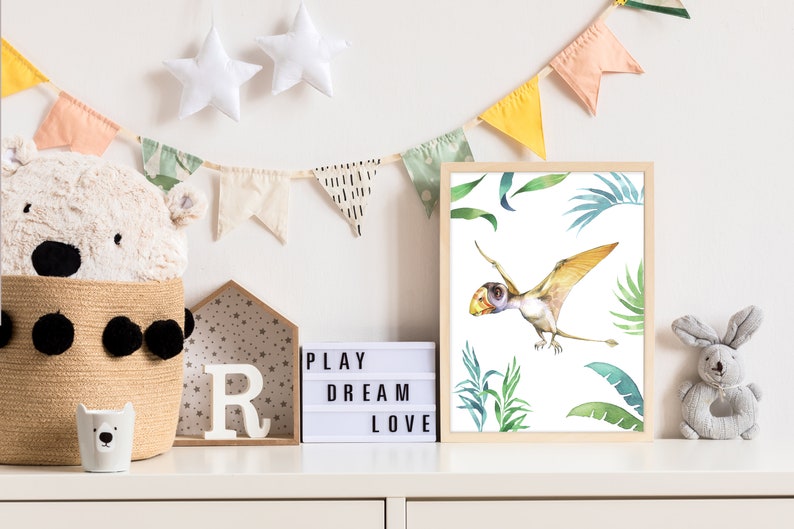 May include: A watercolor illustration of a pterodactyl flying in a jungle setting. The illustration is framed and hanging on a white wall with a light box that reads "PLAY DREAM LOVE" and a stuffed animal rabbit.