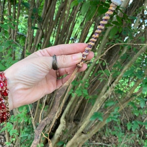 May include: A wooden wand with a skull at the top. The wand is decorated with purple and brown beads. The wand is held in a hand.