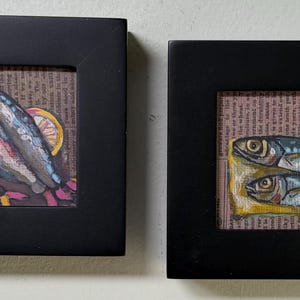 May include: Two framed artworks featuring painted fish and lemon slices on a background of printed text. The left frame contains three fish, while the right frame displays two fish resting on a yellow surface, both signed by the artist Kelly Biren.