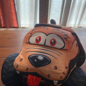 May include: A plush toy shaped like a dog with a cartoon face, orange fur, and a red tongue, featuring large black monster truck tyres. A wooden ruler is positioned vertically behind the toy, showing a height measurement of approximately 12.7 cm.