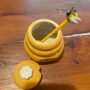 Peut inclure: Un pot à miel en céramique jaune avec un couvercle assorti orné d'un bouton en forme de fleur blanche. Une cuillère à miel jaune avec une figurine d'abeille sur le dessus repose dans le pot. Le pot a une forme arrondie et étagée.