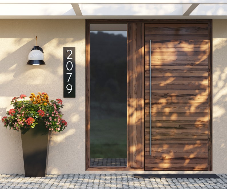 Pu&ograve; includere: Una casa moderna con una porta in legno e una targa con il numero civico nero con numeri bianchi che indicano "2079".
