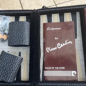 May include: A Pierre Cardin backgammon set in a black textured case. The set includes a brown rule book, two black dice cups, a plastic bag with dice, and a stack of red, black, and cream-colored checkers arranged on the game board surface.