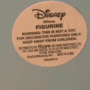 May include: A round sticker with the Disney logo and the word "FIGURINE" in bold. The sticker warns that the item is not a toy and is for decorative purposes only. It also includes the distributor's information and the text "MADE IN CHINA".
