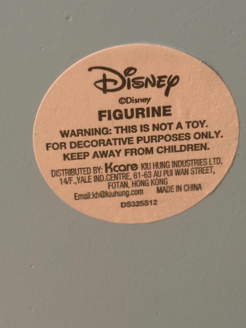 May include: A round sticker with the Disney logo and the word "FIGURINE" in bold. The sticker warns that the item is not a toy and is for decorative purposes only. It also includes the distributor's information and the text "MADE IN CHINA".