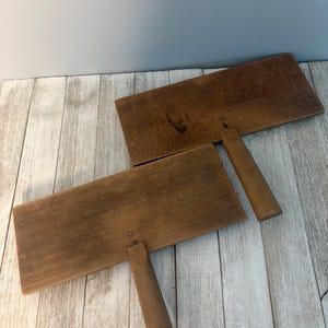 May include: Two rectangular wooden hand cards with brown, weathered surfaces and sturdy handles. Each tool features a flat, elongated wooden base with a centered handle attached by small metal fasteners, showing natural wood grain and wear.
