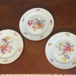 May include: Three white porcelain plates with floral designs. Each plate features a different arrangement of colorful flowers, including pink roses, purple irises, and orange blossoms. The plates have gold rims and are set on a dark brown surface.
