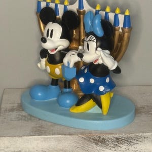 May include: A decorative figurine featuring Mickey and Minnie Mouse standing in front of a gold-colored menorah. Mickey wears yellow shorts and blue shoes, while Minnie dons a blue polka-dot dress and yellow shoes. The base is light blue.